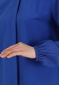 Blusa azul real con puños de mangas fruncidos. Tela suave con un brillo sutil, que presenta un cierre frontal y un ajuste relajado.