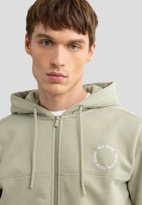 Hellgrüner Reißverschluss-Hoodie mit glatter Textur, Kordelzügen, Seitennähten und einem runden weißen Logo auf der linken Brustseite.