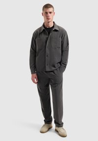 Grijze button-up jas met twee borstzakken en bijpassende grijze broek. Gestyled met een zwart shirt eronder en lichtkleurige schoenen.