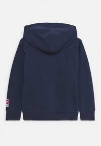 Námořnický modrý svetr s dlouhými rukávy a nášivkou na levém zápěstí, zobrazený ze zadní strany na jednoduchém pozadí.