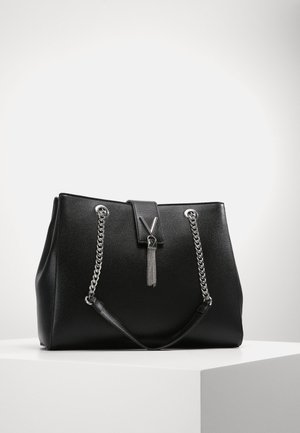 Sac à main en cuir noir avec des bandoulières en chaîne argentées et un fermoir en forme de V avec un pompon argenté, posé sur un socle blanc.