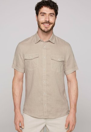 Camisa de botones de manga corta en color beige, hecha de tejido ligero, con dos bolsillos frontales y detalle de botón en la tapeta.