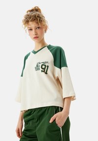 Cremefarvet og grøn kortærmet t-shirt med v-hals, som har "New York 91" broderet i mørkegrøn, og en løs, cropped pasform.
