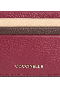 Pouzdro na karty z vínové kůže s embosovanou strukturou, s krémovými a tmavě hnědými vodorovnými pruhy. Zlaté logo "COCCINELLE" ve spodní části.