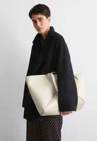 Sac en cuir blanc avec un design replié unique et une longue bandoulière, tenu par une personne en manteau noir, sur fond uni.