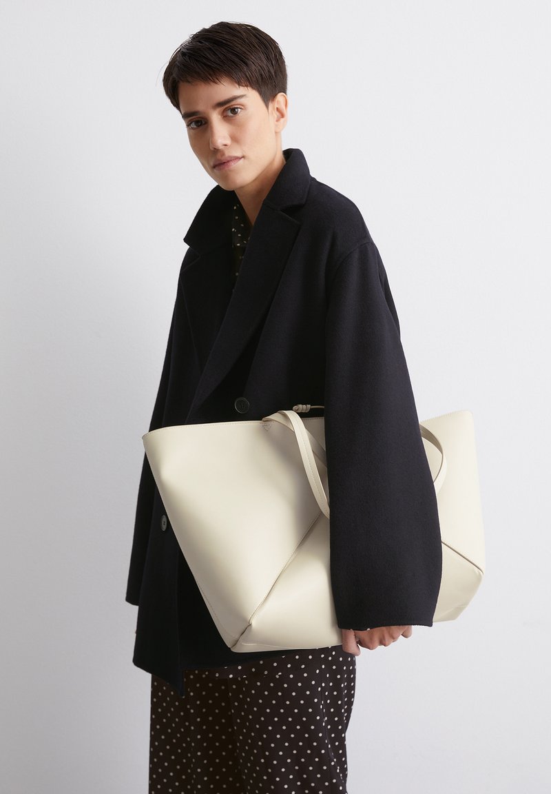 Sac en cuir blanc avec un design replié unique et une longue bandoulière, tenu par une personne en manteau noir, sur fond uni.