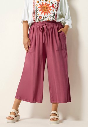 Kobieta nosząca luźne szerokie spodnie w kolorze mauve z wiązanym paskiem w talii, zestawione z białymi sandałami i białym topem z nadrukiem motywu słońca.