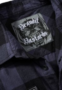 Camicia a quadri nera e blu scuro con etichetta grafica con design di teschio, tag di taglia M e bottoni neri su un tessuto testurizzato.
