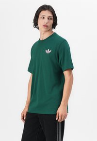 Zaļa kokvilnas T-krekls ar apaļu dekoltē, īsām piedurknēm un rozā Adidas logotipu kreisajā krūtis pusē. Uzvilkts kopā ar tumšiem džinsiem.