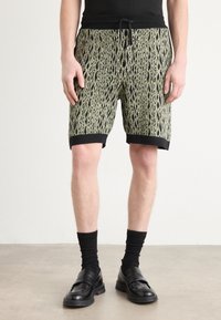 Pantalones cortos para hombre de un tejido verde texturizado con un patrón de serpiente, con una cinturilla y ribetes negros, combinados con calcetines negros y mocasines.