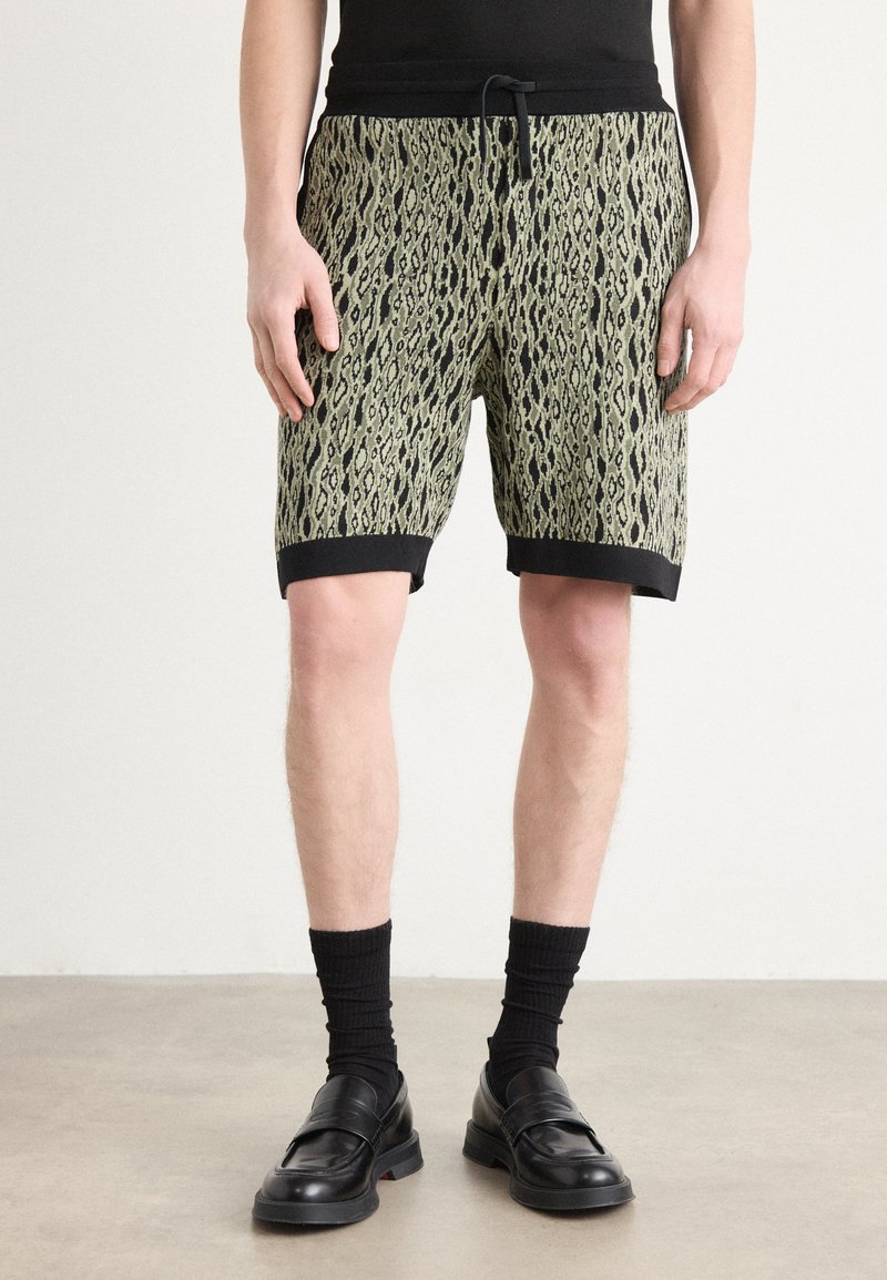 Pantalones cortos para hombre de un tejido verde texturizado con un patrón de serpiente, con una cinturilla y ribetes negros, combinados con calcetines negros y mocasines.