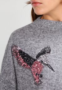 Un gros plan d'une femme portant un pull léger gris avec un motif de colibri en sequins noirs et roses sur la zone de la poitrine.