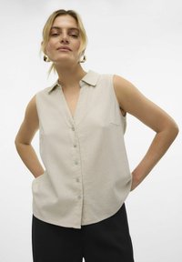 Blouse sans manches beige clair avec col, dotée d'un devant à boutons et de poches latérales, fabriquée dans un tissu doux.
