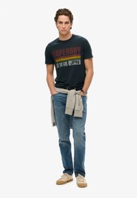 Mørkeblå T-shirt med teksten "SUPERDRY" og "JPN" i rød, gul og hvid. Lyseblå jeans, lys grå sweatshirt bundet om livet, beige sneakers.