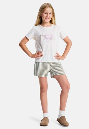 Lachend meisje staat met handen in de zij, draagt een wit t-shirt met een roze hart en bloemen, gestreepte short, witte sokken en bruine instapschoenen.