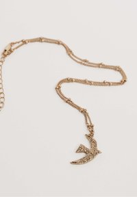 Arany tónusú nyaklánc, madár alakú medállal, amelynek felülete textúrált, finom lánchoz csatlakozik, apró gyöngy díszítéssel.