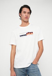 Young man with dark hair wearing a white Tommy Hilfiger T-shirt and blue jeans, standing with one hand in pocket against a plain background.