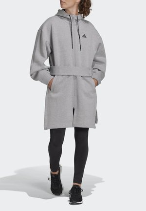 Woman wearing a long grey zip-up hoodie with a belt, black leggings, and black sneakers, walking with hands in pockets.