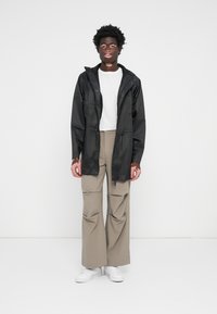 Veste imperméable noire avec capuche ; pantalon long gris ; t-shirt blanc ; le modèle se tient sur un fond neutre. Les chaussures sont blanches, avec un design minimal.