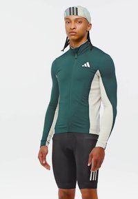 Atletisk cykeloutfit med en långärmad tröja i mörkgrönt med en krämfärgad panel och svarta shorts, prydda med vita ränder och logotyper.