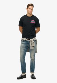 Černé bavlněné tričko s barevným designem hor a nápisem "Superdry". Nosit se světle modrými džíny a šedým svetrem uvázaným kolem pasu.