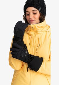 Moufles noires imperméables avec des sangles ajustables, design rembourré et accents de logo. Portées par une personne en veste jaune et bonnet noir.