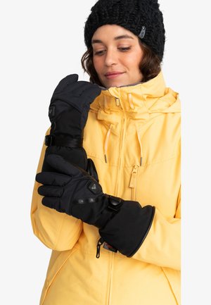 Moufles noires imperméables avec des sangles ajustables, design rembourré et accents de logo. Portées par une personne en veste jaune et bonnet noir.