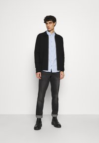 Svart genser med glidelås over en lyseblå skjorte, kombinert med mørke jeans og svarte støvler, plassert mot en hvit bakgrunn.