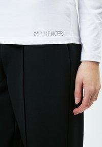 Weißes Langarmhemd mit "INFLUENCER" in Strass auf der Seite, kombiniert mit schwarzen maßgeschneiderten Hosen. Weicher Stoff, lockere Passform.