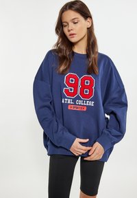Felpa oversize blu navy con testo ricamato bianco e rosso che recita "ATHL. COLLEGE 98 SPORTS." Maniche lunghe e vestibilità rilassata. Leggings neri.