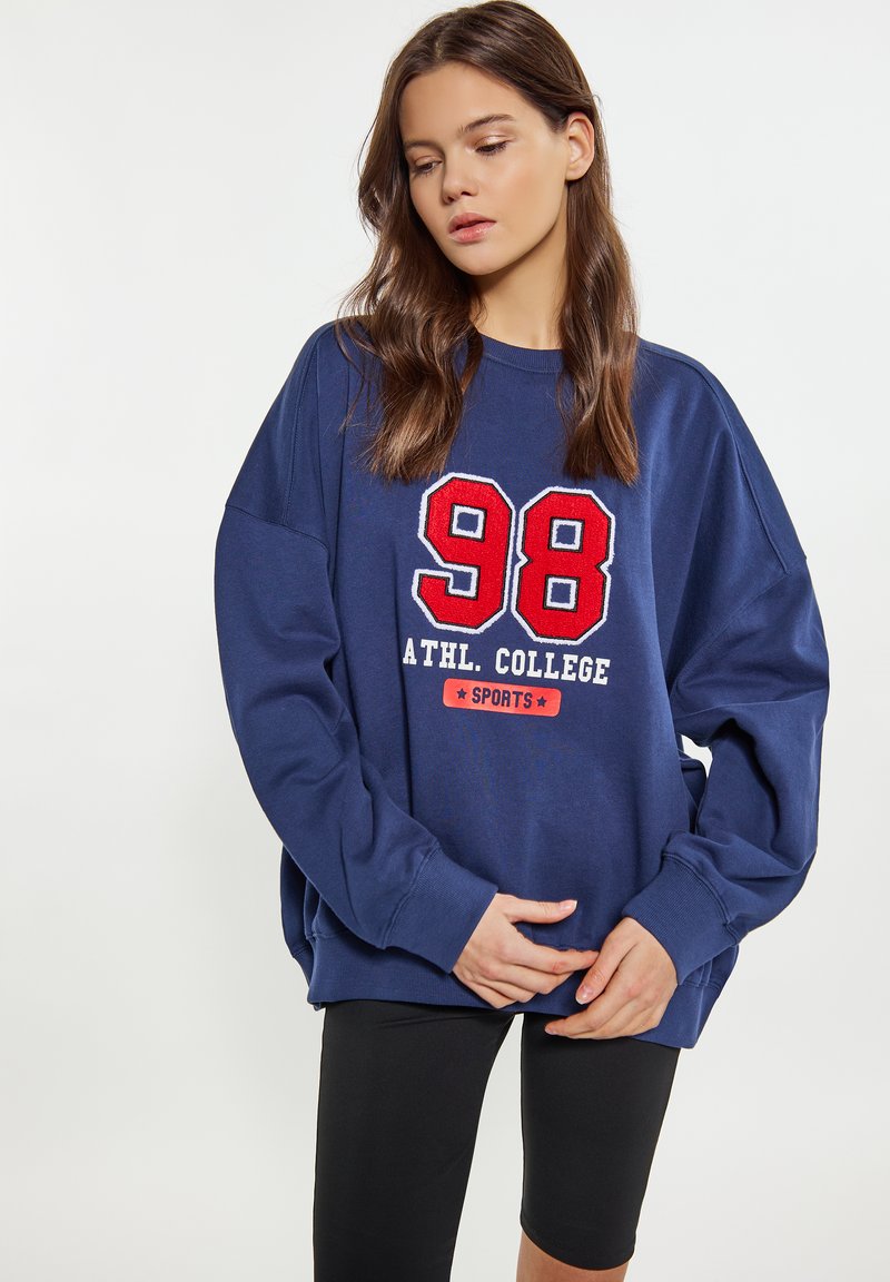 Felpa oversize blu navy con testo ricamato bianco e rosso che recita "ATHL. COLLEGE 98 SPORTS." Maniche lunghe e vestibilità rilassata. Leggings neri.