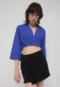 Camicia blu corta con scollatura profonda e maniche larghe abbinata a una gonna nera avvolgente con dettaglio decorativo a nodo.