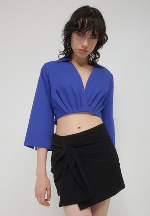Camicia blu corta con scollatura profonda e maniche larghe abbinata a una gonna nera avvolgente con dettaglio decorativo a nodo.