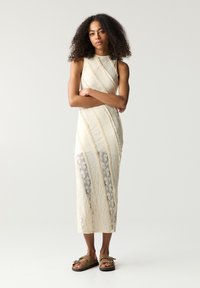 Krepp-Midi-Kleid in Creme mit diagonalen strukturierten Streifen und transparenten Einsätzen. Kombiniert mit braunen Sandalen, die mit Metallakzenten verziert sind.