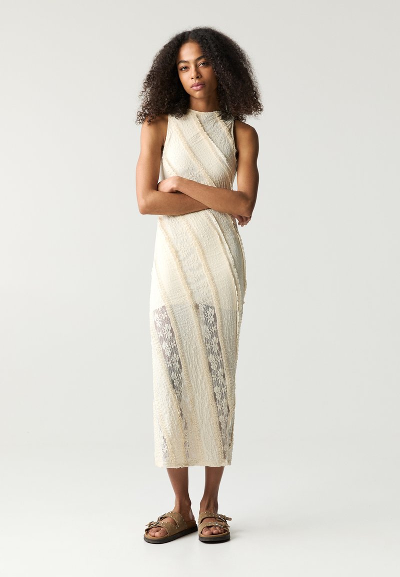 Krepp-Midi-Kleid in Creme mit diagonalen strukturierten Streifen und transparenten Einsätzen. Kombiniert mit braunen Sandalen, die mit Metallakzenten verziert sind.