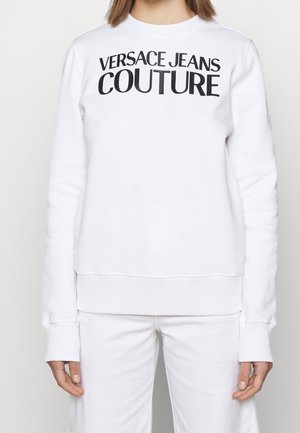 Person iført en hvid sweatshirt og bukser, foran på sweatshirt står der "VERSACE JEANS COUTURE" med sort tekst.