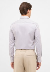 Langärmliges Hemd in helllila mit kleinen, dunklen Rautenmustern. Es hat einen spitzen Kragen und geknöpfte Manschetten, kombiniert mit beigen Hosen.