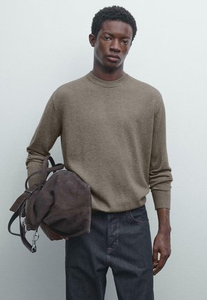 Light brown knitted sweater with a round neckline and ribbed cuffs, paired with dark denim jeans. Model holds a dark brown suede bag.