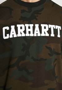 Camo-trui in aardetinten met een geribbelde zwarte halslijn en opvallende witte "CARHARTT"-letters over de borst. Zachte katoenen textuur.