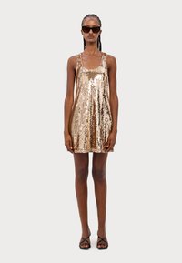 Robe mini en sequins dorés avec un décolleté arrondi et des bretelles spaghetti, présentant une coupe ample et une texture brillante. Modèle portant des sandales noires à lanières.
