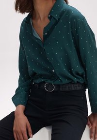 Camicia verde scuro con piccoli motivi turchesi, design a bottoni, maniche arrotolate, abbinata a pantaloni neri e una cintura intrecciata con una fibbia rotonda.