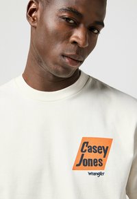 Sudadera de algodón blanca con un gráfico en naranja y negro que incluye el texto "Casey Jones" y "Wrangler". Cuello redondo y textura suave.