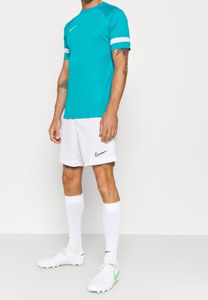 Homme portant un t-shirt de sport turquoise à manches courtes, un short blanc, de longues chaussettes blanches et des chaussures à crampons blanches avec des détails verts, debout sur fond blanc.