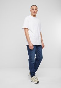 Camiseta blanca de algodón, corte relajado, combinada con jeans de mezclilla azul. Modelo lleva zapatillas beige con acentos grises. Fondo neutro.