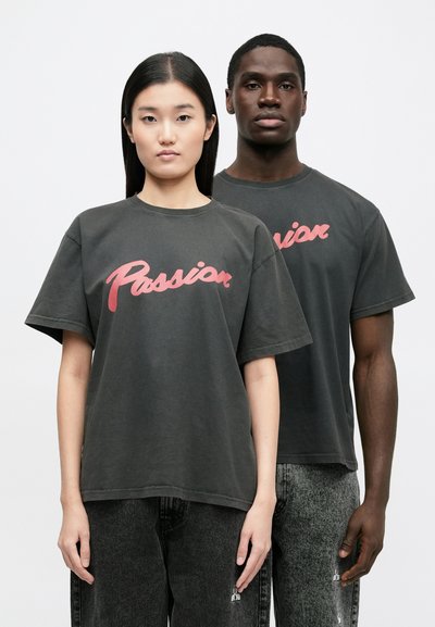 Grå oversizade t-shirts med "Passion" i rosa skrivstil. Bomullsmaterial, rund hals, korta ärmar. Modellen bär mörkgrå jeans.