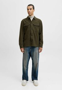Mand står iført en olivengrøn knap-uppet skjorte over en beige højhalset trøje, slidte løse jeans og hvide sneakers foran en ensfarvet hvid baggrund.