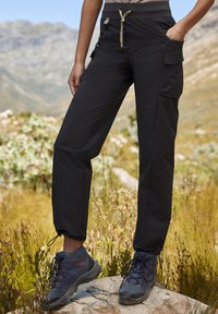 Schwarze Outdoor-Hosen mit elastischem Bund und Kordelzug, ausgestattet mit seitlichen Taschen und schmal zulaufenden Bündchen, kombiniert mit dunklen Wanderschuhen.