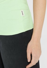 Helle grüne Baumwollbluse mit tailliertem Design, versehen mit einem kleinen weißen "SENSES"-Label an der Seite. Kombiniert mit dunklen Jeans.