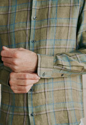 Man adjusting cuff of green plaid long-sleeve button-up shirt with blue and beige lines.