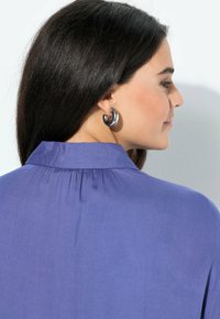 Blusa viola con colletto e dettagli plissettati sul retro, abbinata a grandi orecchini rotondi in argento. Tessuto a trama liscia.